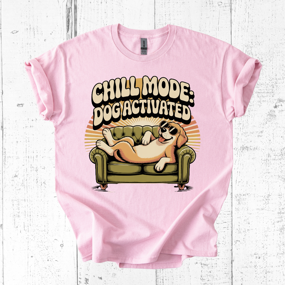 Pink t-shirt with a graphic of a dog on a couch and text 'Chill Mode: Dogiactivated' on a white wooden background.