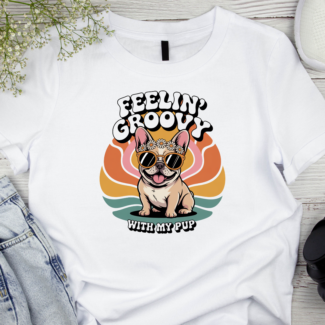 White t-shirt with a dog graphic and 'Feelin' Groovy with my pup' text on a wooden surface.