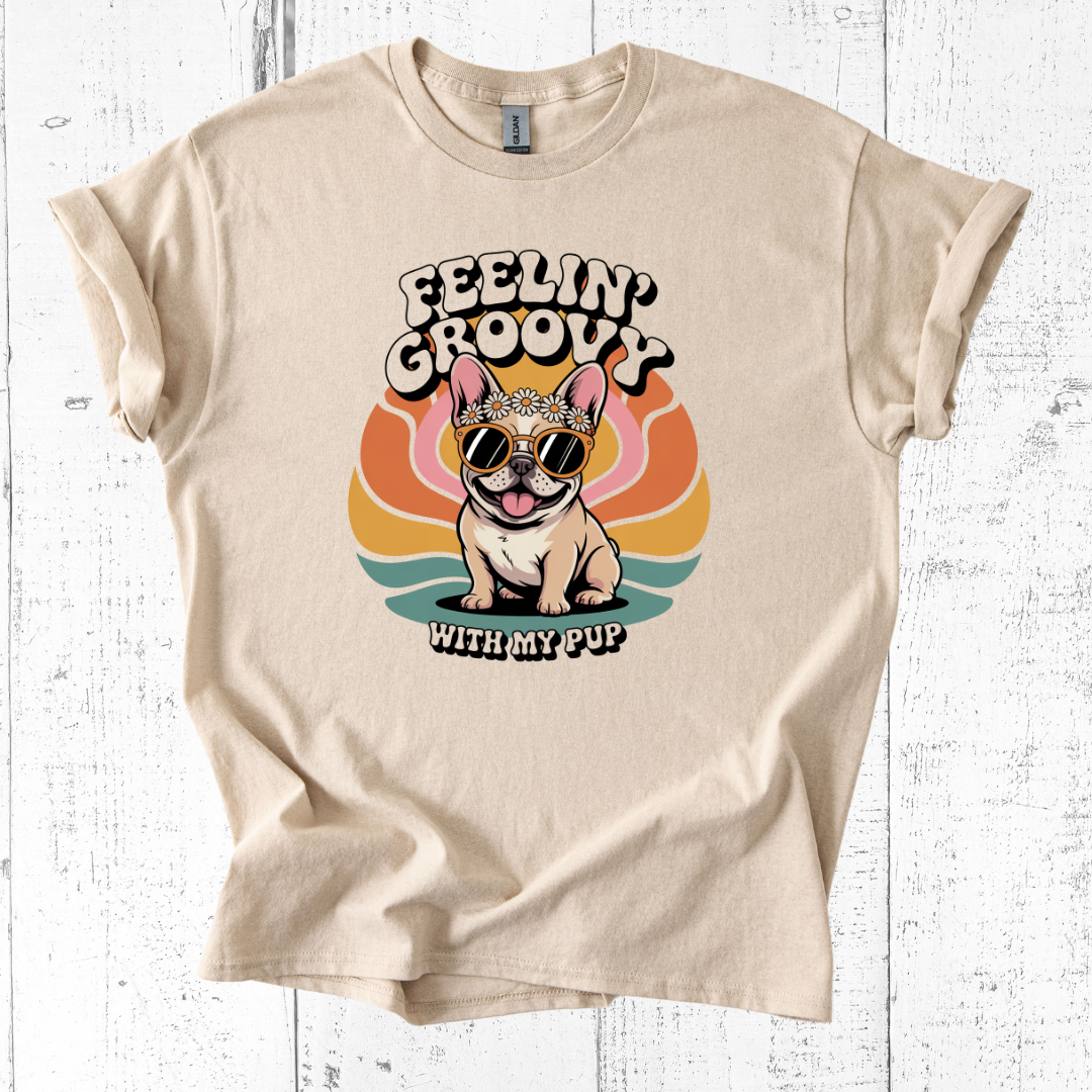 Beige t-shirt with a graphic of a dog wearing sunglasses and a bandana, with text 'Feelin' Groovy with my pup' on a white wooden background.