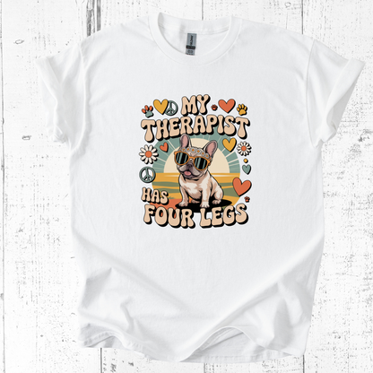 White t-shirt with a dog graphic and text 'My therapist has four legs' on a wooden surface.