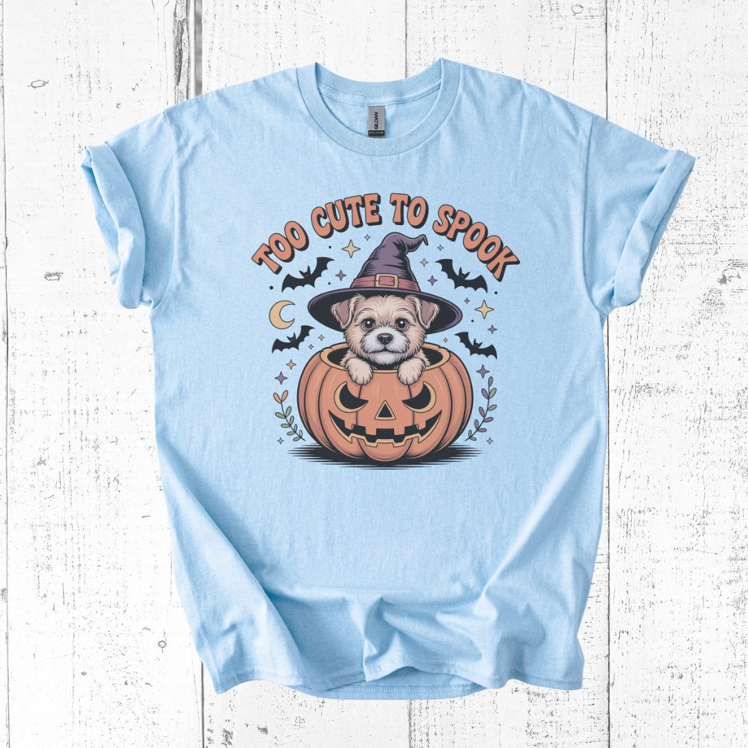 Light blue t-shirt with a Halloween-themed design of a dog in a pumpkin and text 'Too Cute to Spook' on a wooden surface.