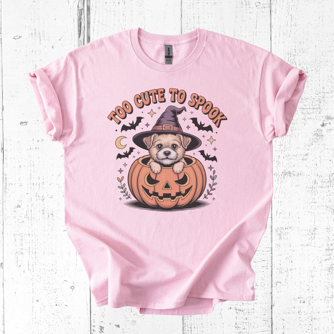 Pink t-shirt with a Halloween design of a dog in a pumpkin and text 'Too Cute to Spook' on a wooden surface.