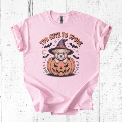 Pink t-shirt with a Halloween design of a dog in a pumpkin and text 'Too Cute to Spook' on a wooden surface.