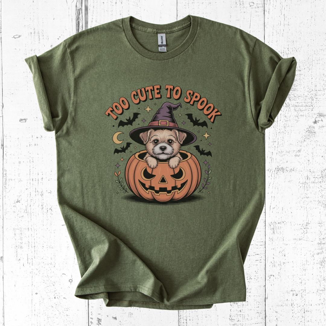 Green t-shirt with a Halloween-themed design of a dog in a pumpkin, text 'Too Cute to Spook', on a white wooden background.
