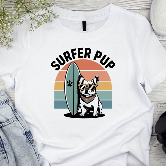 White t-shirt with 'Surfer Pup' design featuring a dog with sunglasses and a surfboard, on a wooden surface.
