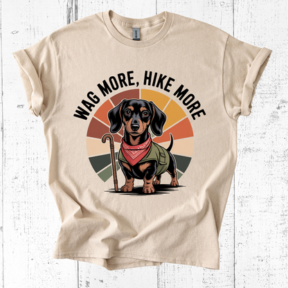 Beige t-shirt with a dachshund graphic and 'Wag More, Hike More' text on a white wooden background