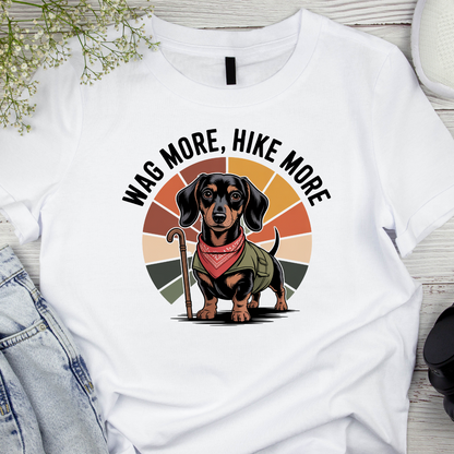 White t-shirt with a dachshund graphic and 'Wag More, Hike More' text on a wooden surface.