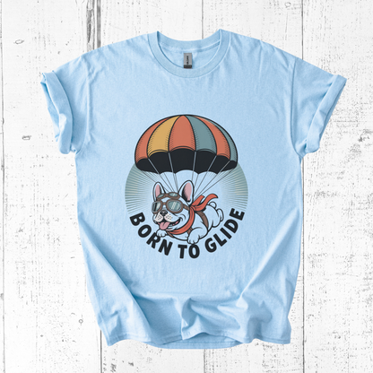 Light blue t-shirt with a graphic of a dog with goggles and a parachute, and the text 'Born to Glide' on a white wooden background.