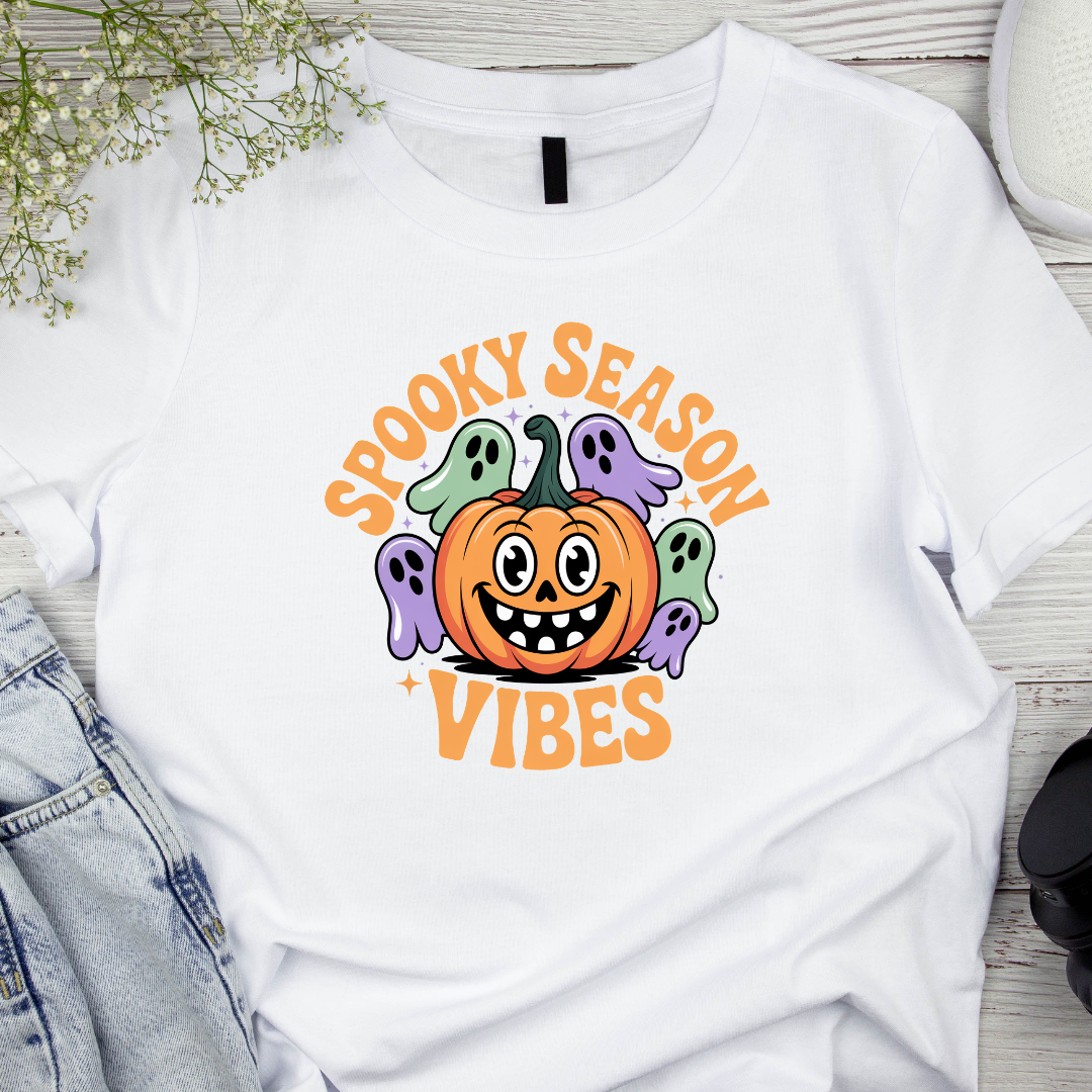 White t-shirt with Halloween-themed design featuring a pumpkin and ghosts on a wooden surface.