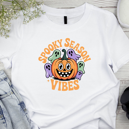 White t-shirt with Halloween-themed design featuring a pumpkin and ghosts on a wooden surface.