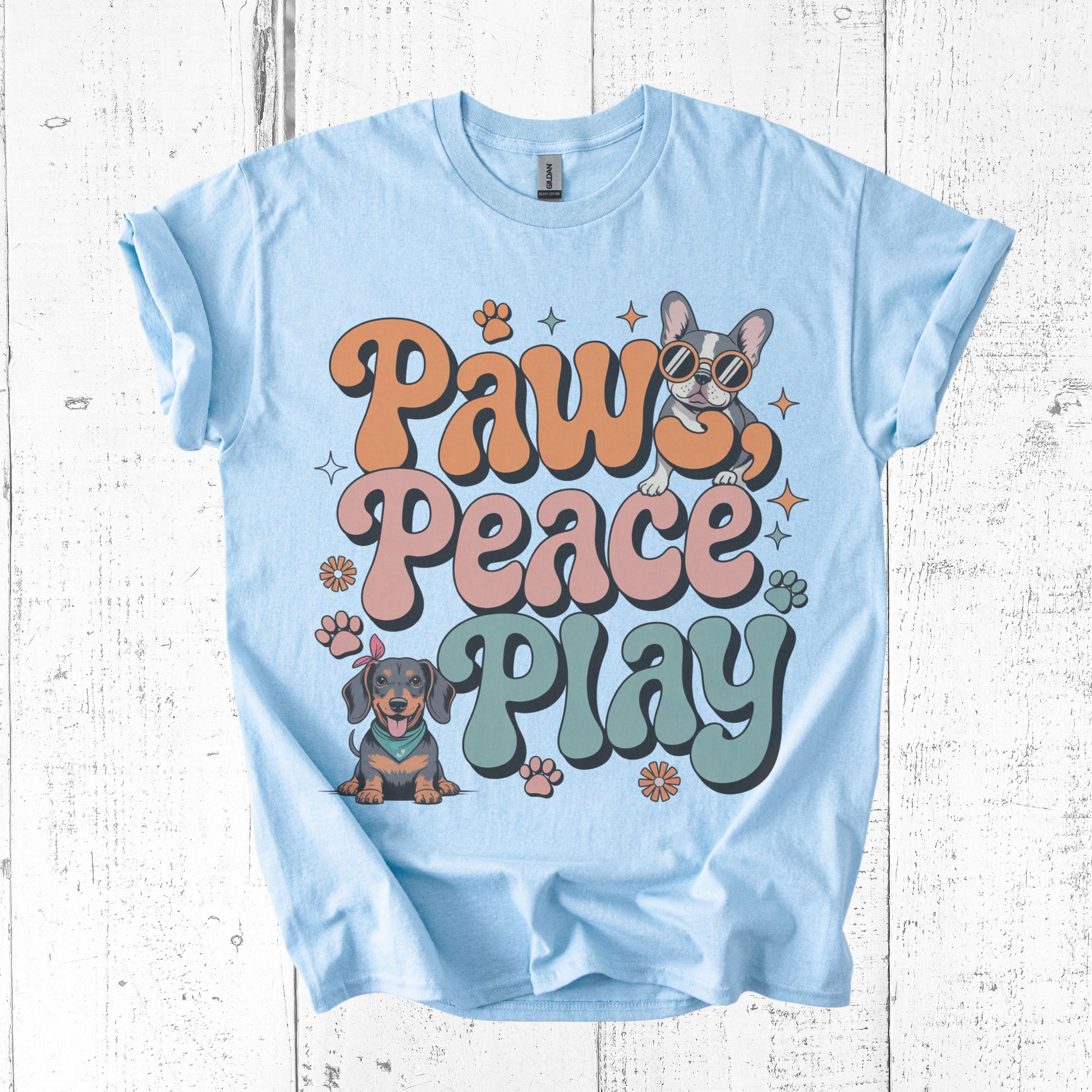 Light blue t-shirt with 'Paws Peace Play' text and dog illustrations on a wooden surface