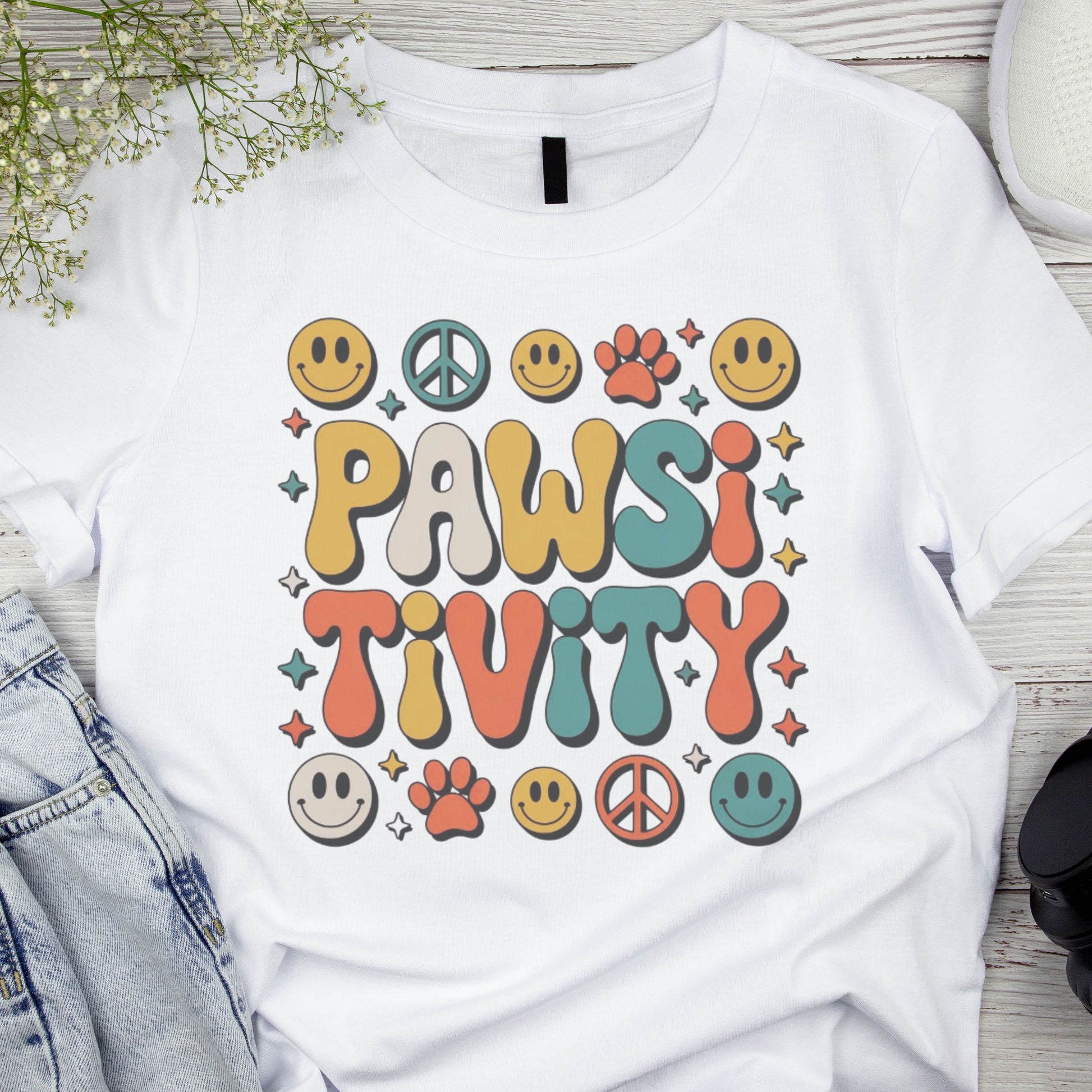 White t-shirt with 'PAWSI TIVITY' design on a wooden surface with denim jeans and flowers.