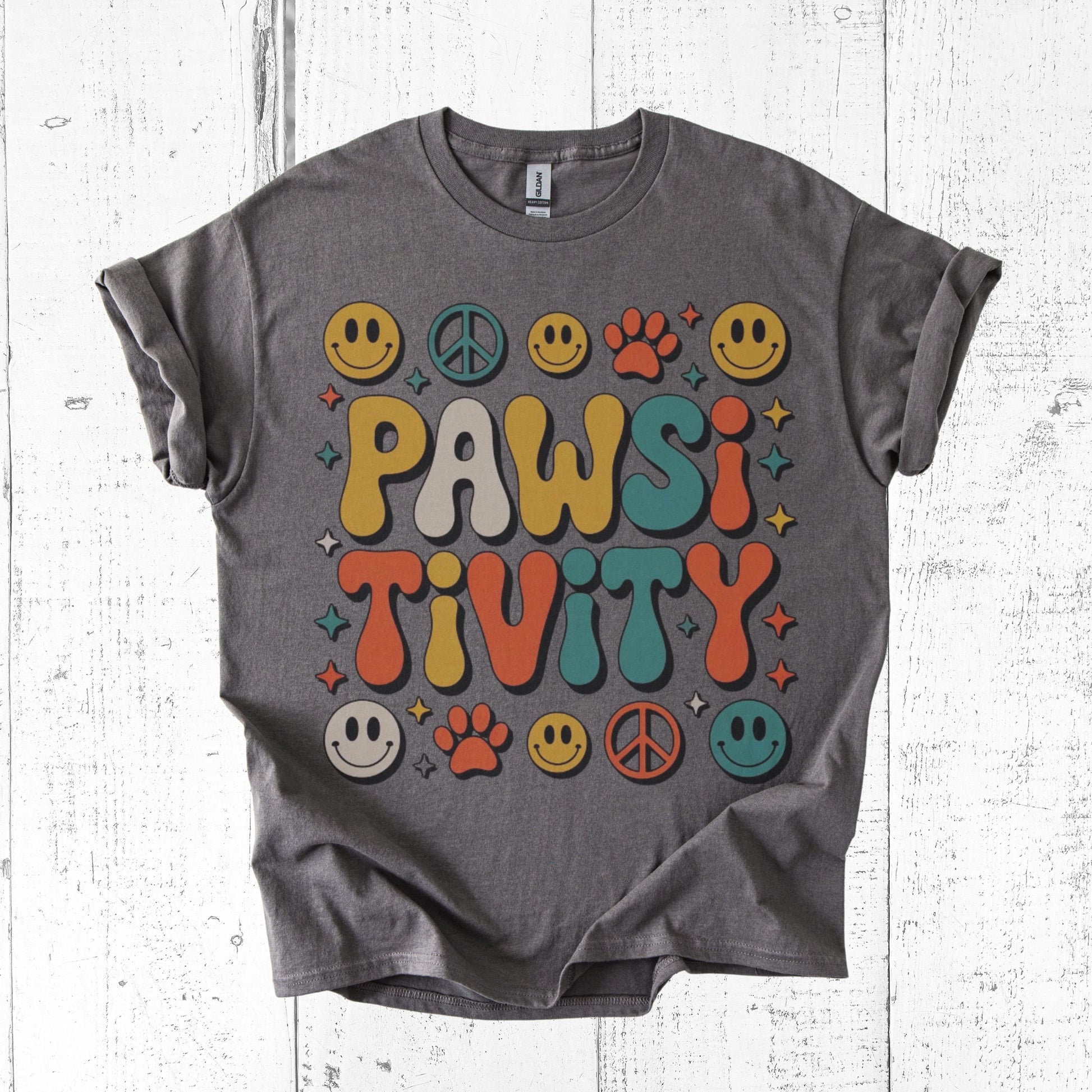 Gray t-shirt with colorful 'PAWSI TIVITY' design on a white wooden surface
