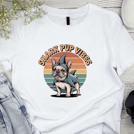 White t-shirt with a dog in a shark costume graphic and 'Shark Pup Vibes' text on a wooden surface.