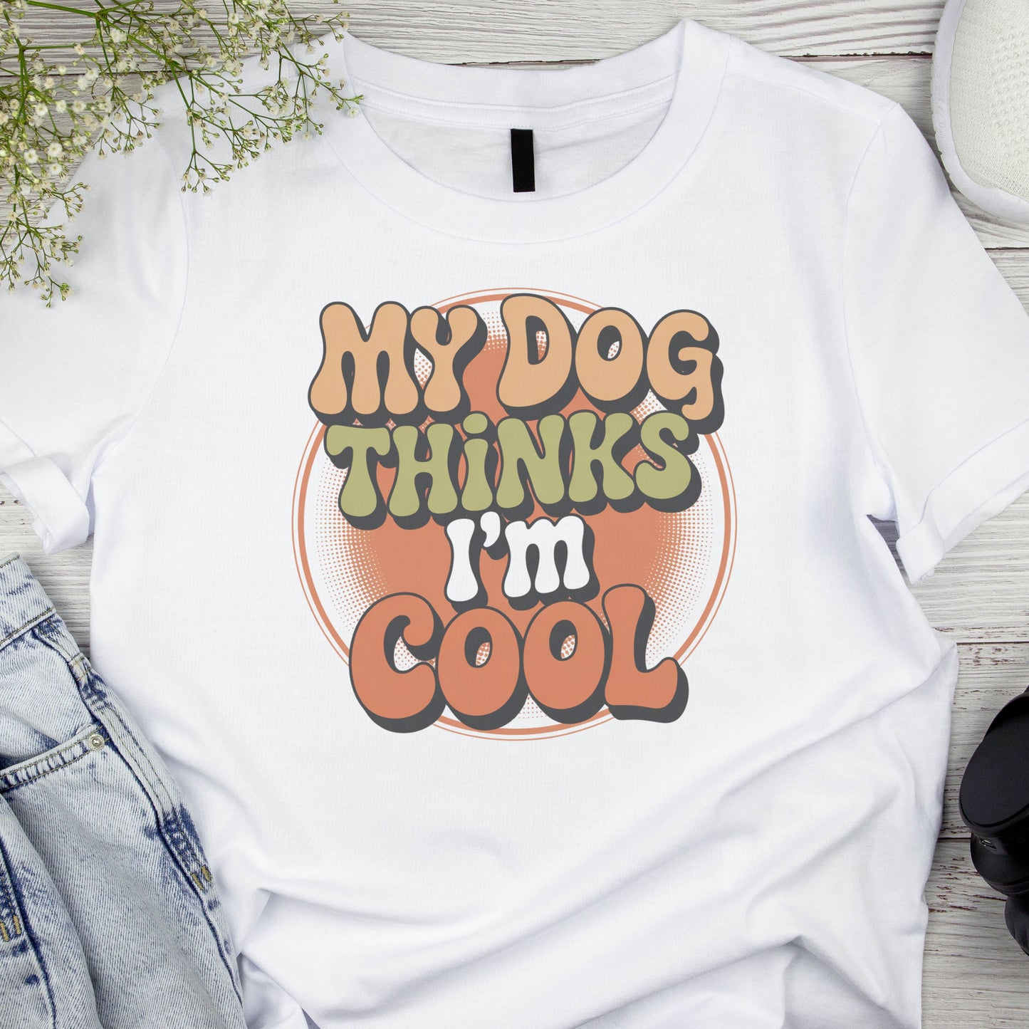 White t-shirt with 'My Dog Thinks I'm Cool' text on a wooden surface with denim jeans and a plant.