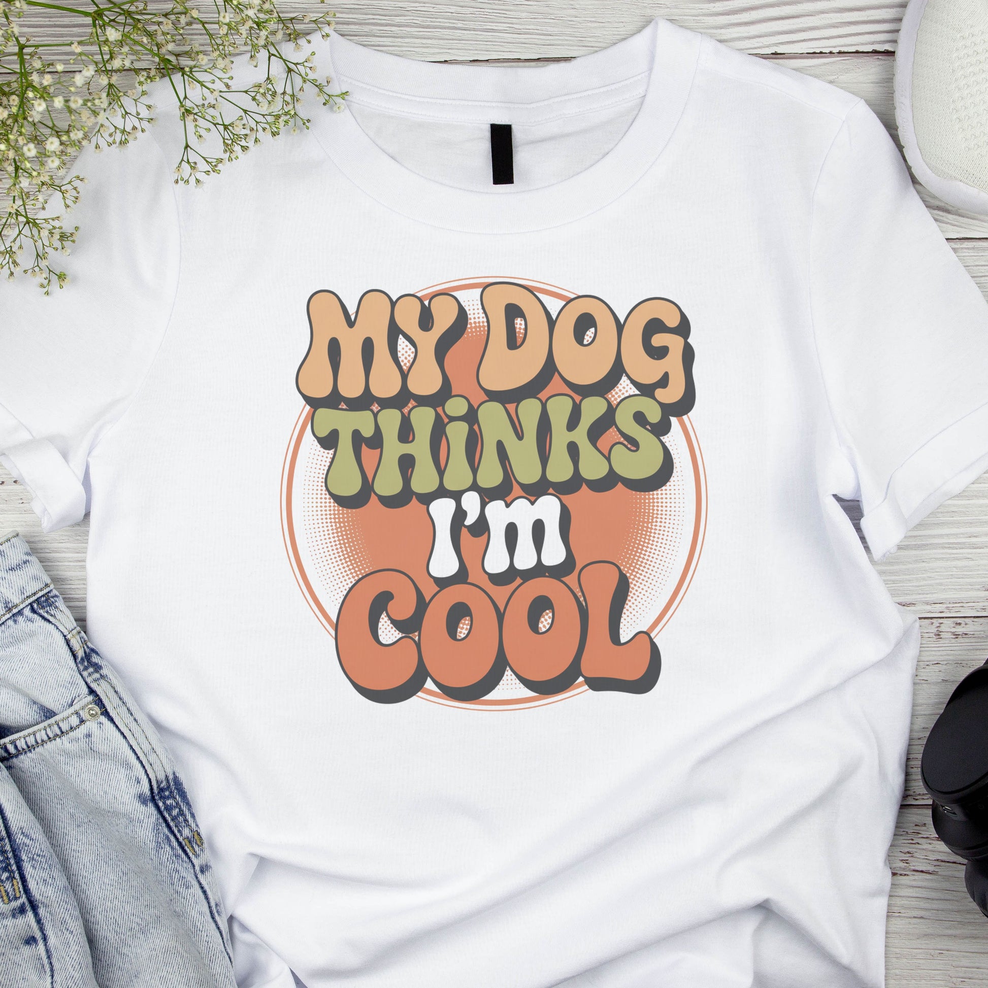 White t-shirt with 'My Dog Thinks I'm Cool' text on a wooden surface with denim jeans and a plant.