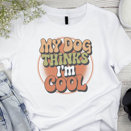 White t-shirt with 'My Dog Thinks I'm Cool' text on a wooden surface with denim jeans and a plant.