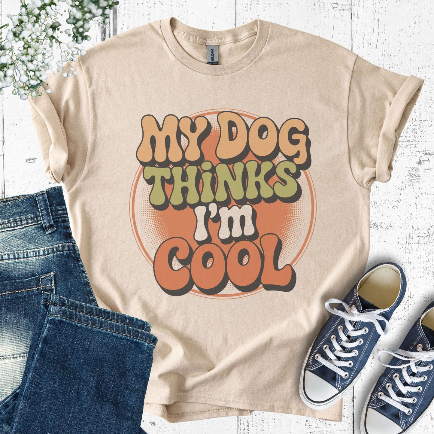 Beige t-shirt with 'My Dog Thinks I'm Cool' text, paired with blue jeans and sneakers on a white wooden surface.