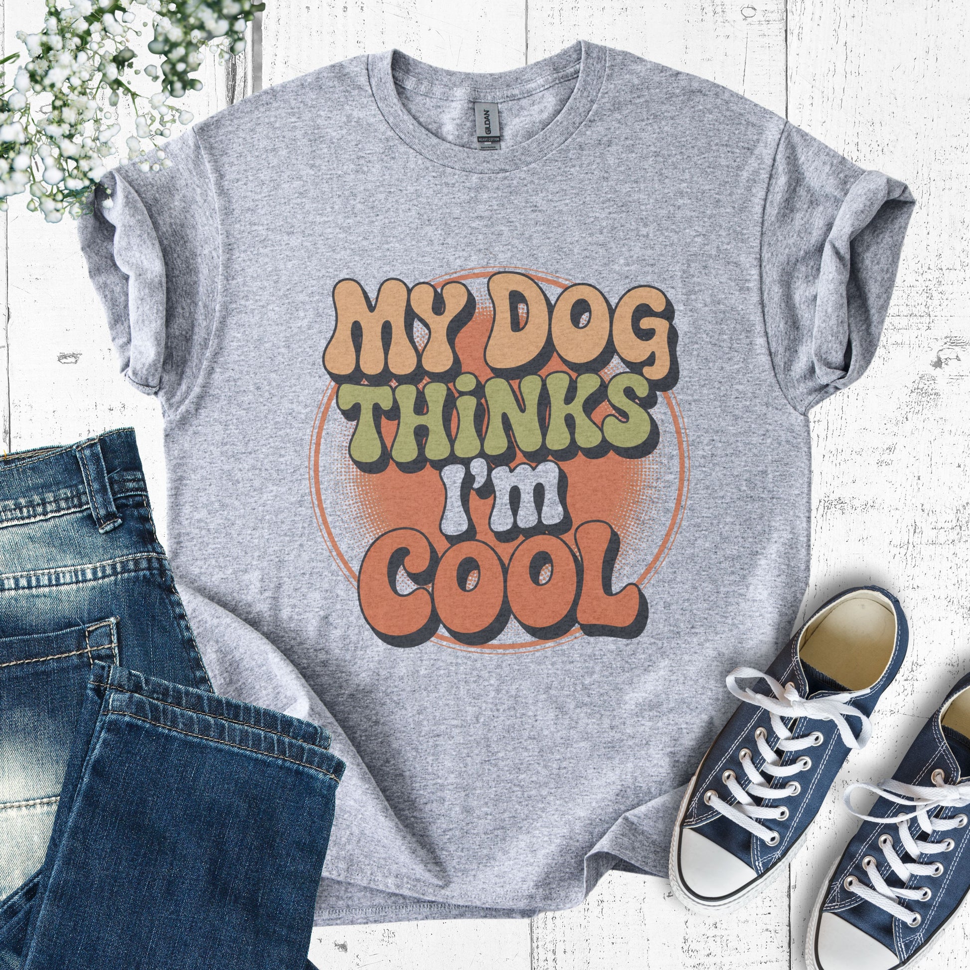 Gray t-shirt with 'My Dog Thinks I'm Cool' text, paired with jeans and sneakers on a light wooden surface.