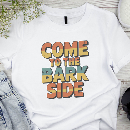White t-shirt with 'Come to the Bark Side' text on a wooden surface with denim jeans and flowers.