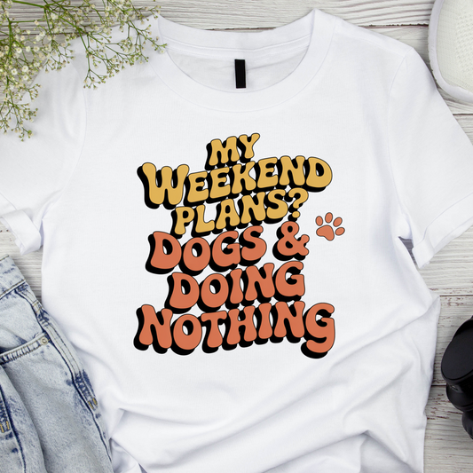 White t-shirt with 'My weekend plans? Dogs & doing nothing' text on a wooden surface.