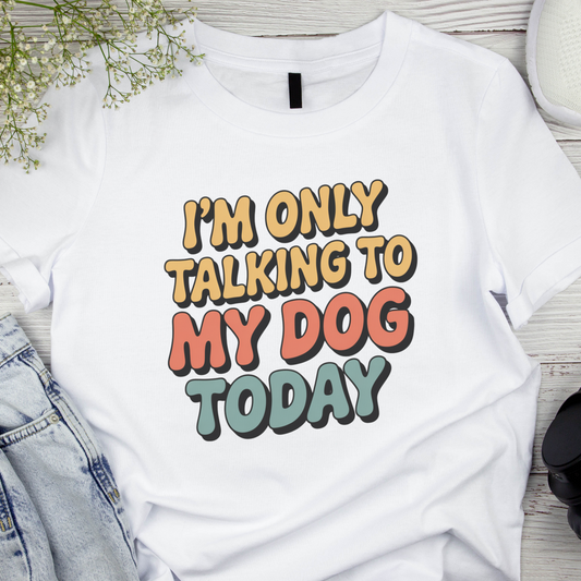 White t-shirt with colorful text 'I'm only talking to my dog today' on a wooden surface.