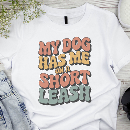 White t-shirt with colorful text 'My dog has me on a short leash' on a wooden surface.