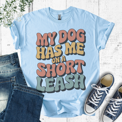 Light blue t-shirt with colorful text 'My dog has me on a short leash' on a wooden surface with jeans and sneakers.