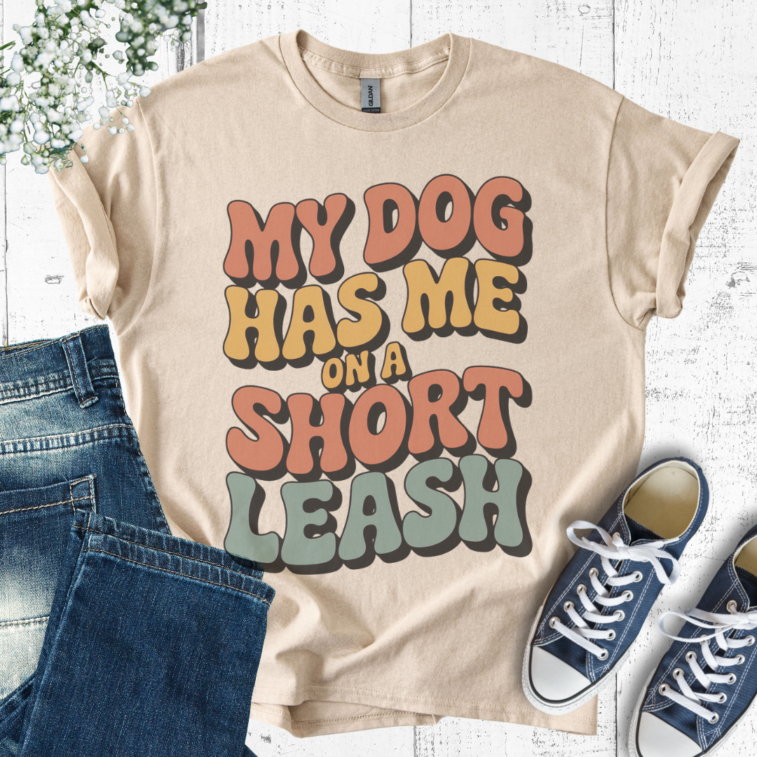 Beige t-shirt with 'My dog has me on a short leash' text, paired with jeans and sneakers on a light wooden surface.