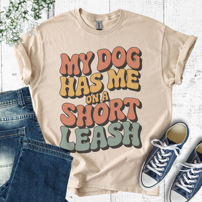 Beige t-shirt with 'My dog has me on a short leash' text, paired with jeans and sneakers on a light wooden surface.