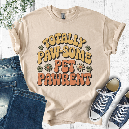 Beige t-shirt with 'Totally Paw-some Pet Pawrent' text, blue jeans, and sneakers on a white wooden surface.