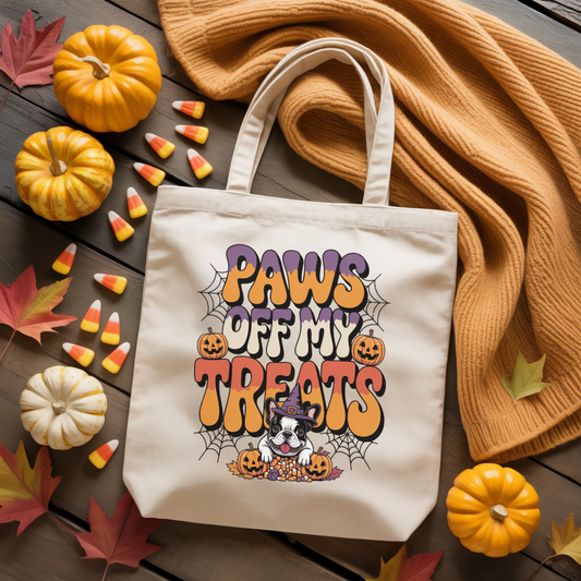 Tote bag with 'Paws Off My Treats' design surrounded by pumpkins, candy corn, and leaves on a wooden surface.