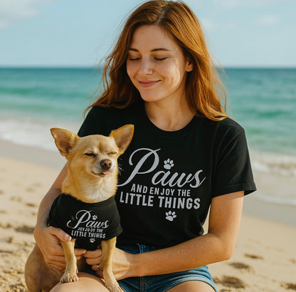 Woman and dog wearing matching 'Paws and enjoy the little things' shirts on a beach.