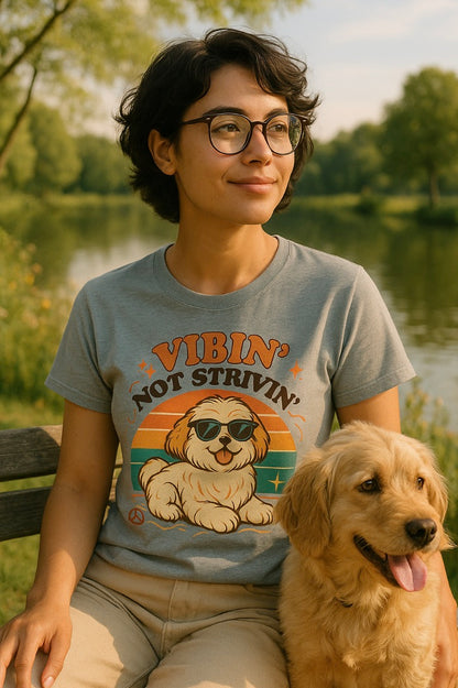 Person wearing a t-shirt with a dog graphic and text, sitting next to a dog by a lake. 'Vibin' Not Strivin'.